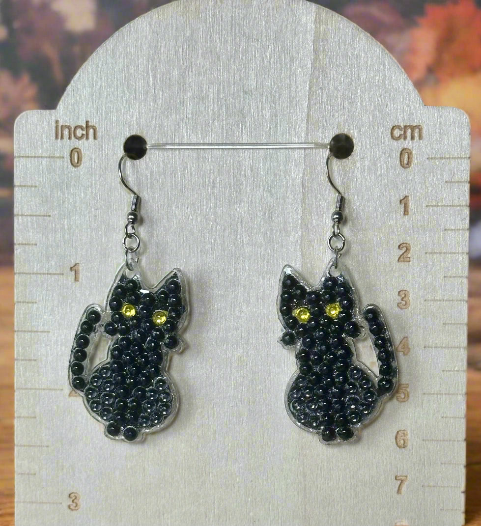 Black cat-shaped earrings with yellow eyes on a white stand with measurement scale.