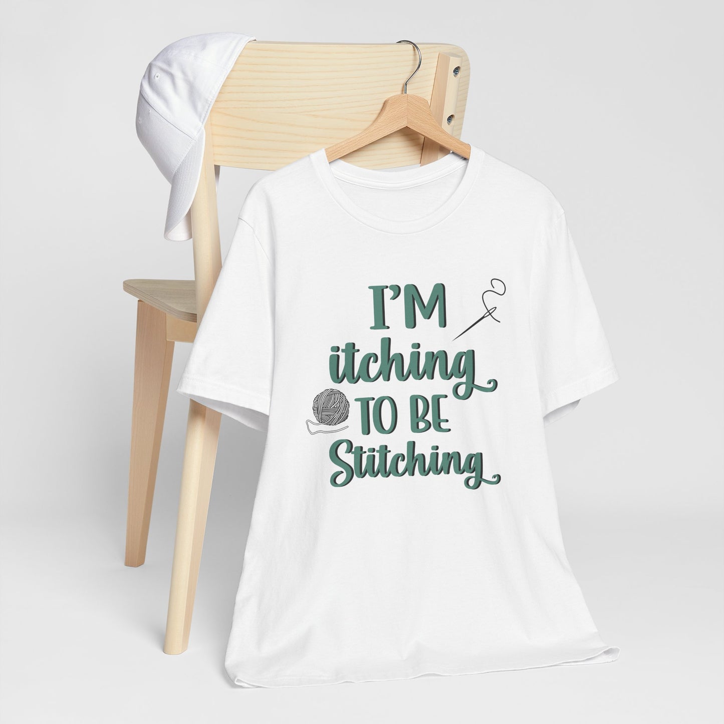 Funny Itching to be Stitching white colored T-Shirt with heather teal lettering, a ball of yarn, and a needle with thread