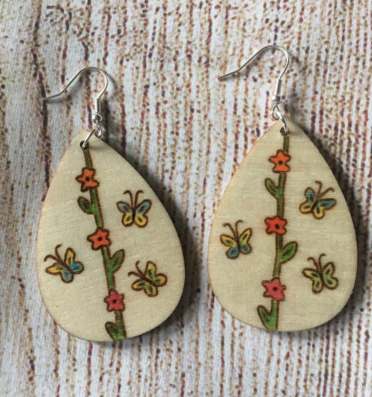 Wood burned earrings with flower vine in the middle from the top to the bottom of the earrings.  red flowers are on the vine with yellow and blue butterflies on either side. Earrings are tear drop shaped with fishhooks.