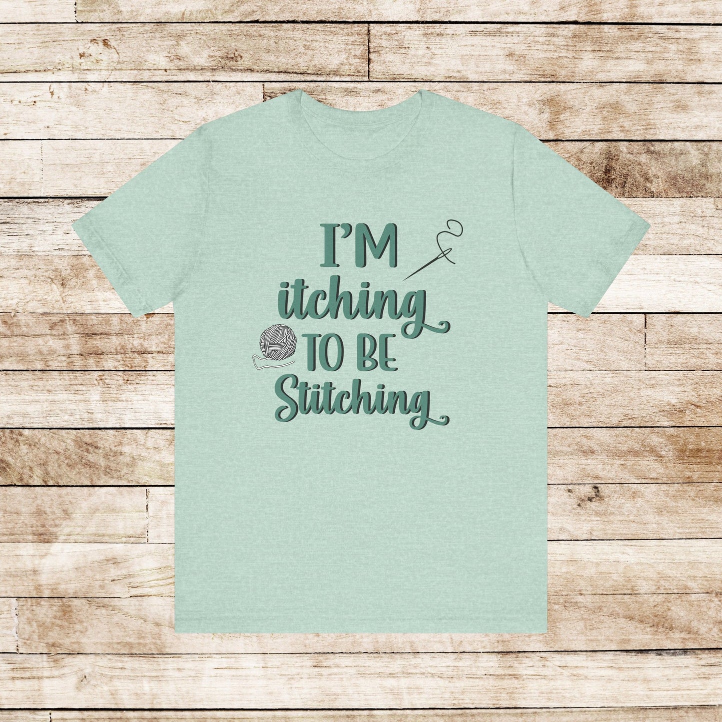 Funny Itching to be Stitching heather prism mint colored T-Shirt with heather teal lettering, a ball of yarn, and a needle with thread