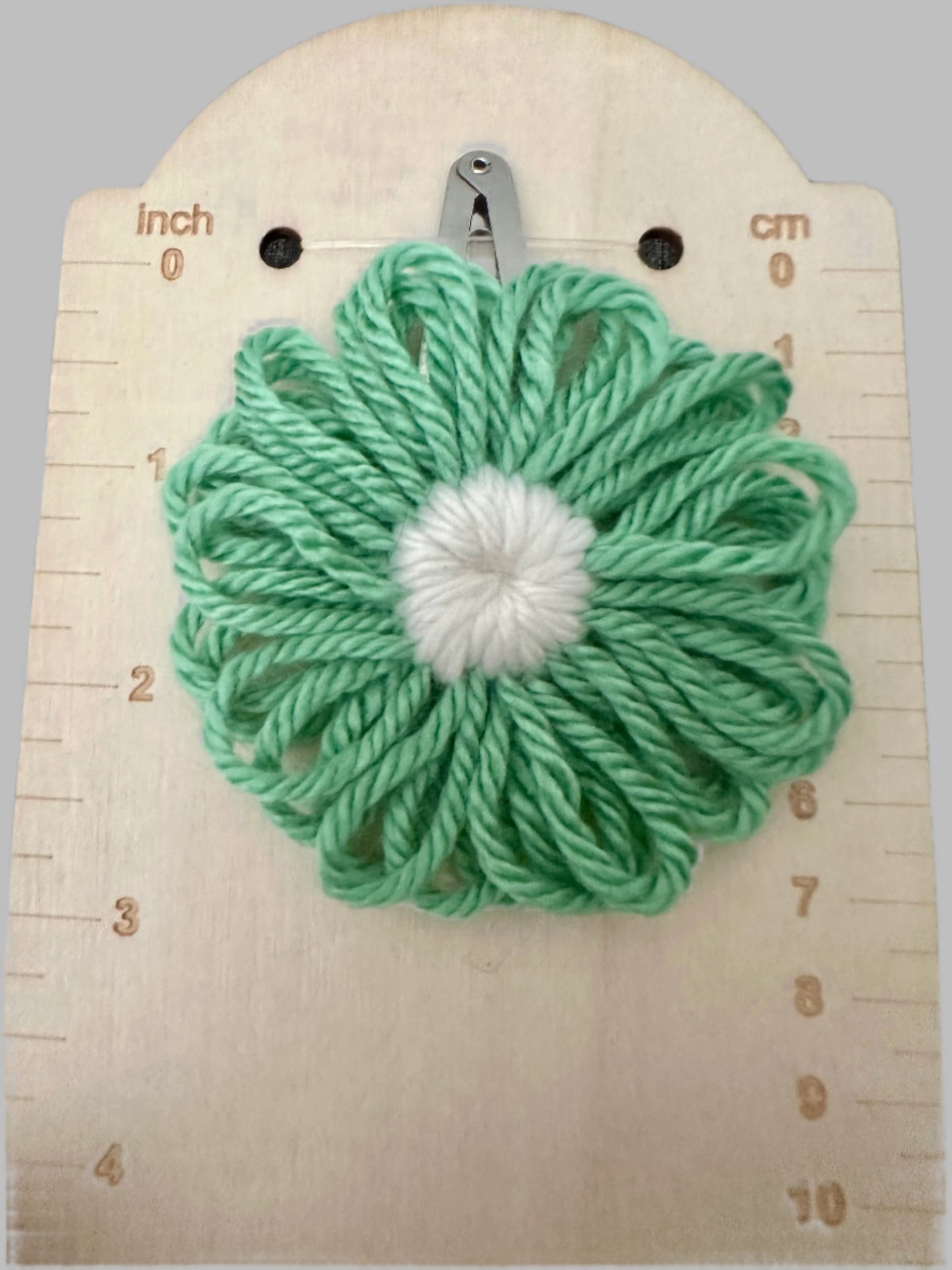 Green yarn flower with a white center on a wooden surface with measurement markings.