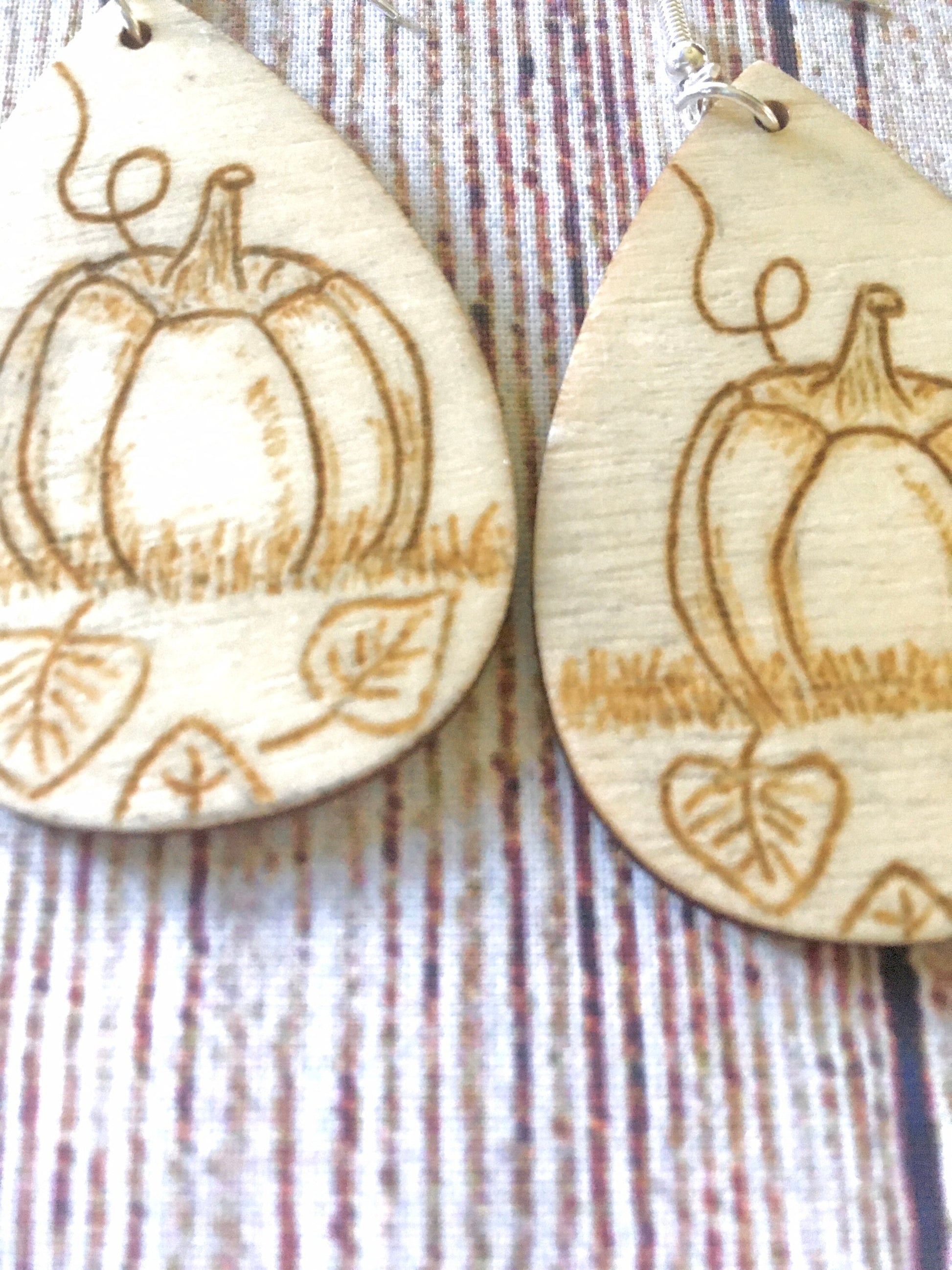 Wood burned earrings with wood burned pumpkin sitting in the grass with leaves on the ground. Earrings are tear drop shaped with fishhooks.