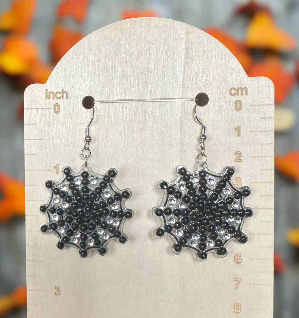 Spider web earrings on a wooden display card with measurement scale, blurred Halloween decorations in the background.