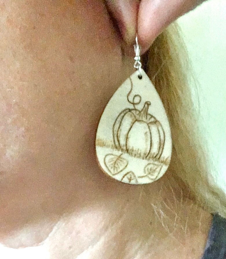 Wood burned earrings with wood burned pumpkin sitting in the grass with leaves on the ground. Earrings are tear drop shaped with fishhooks.
