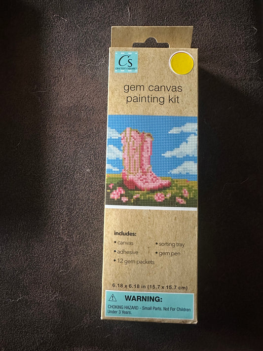 Cardboard packaging of a gem canvas painting kit on a dark surface.   Kit is pink cowgirl boots on grass with pink flowers, with blue sky with clouds