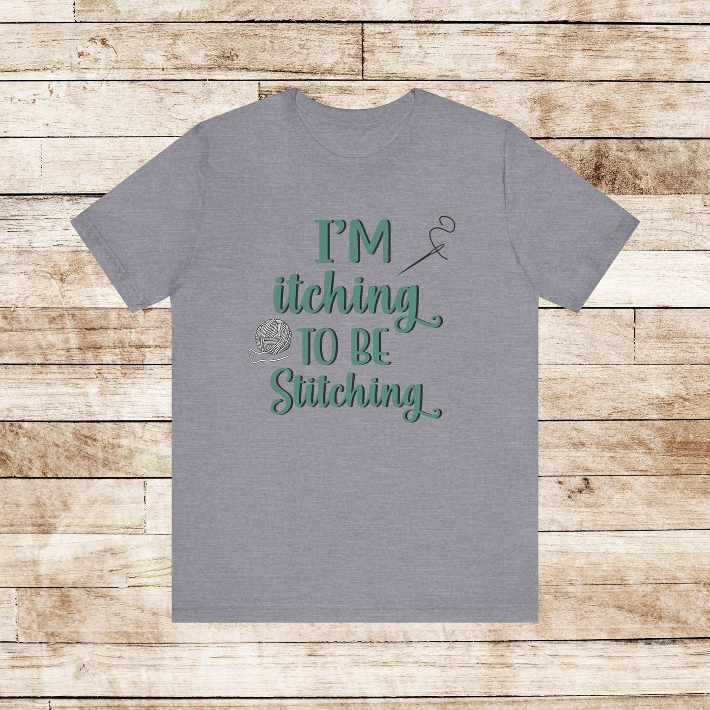 Funny Itching to be Stitching heather storm colored T-Shirt with heather teal lettering, a ball of yarn, and a needle with thread
