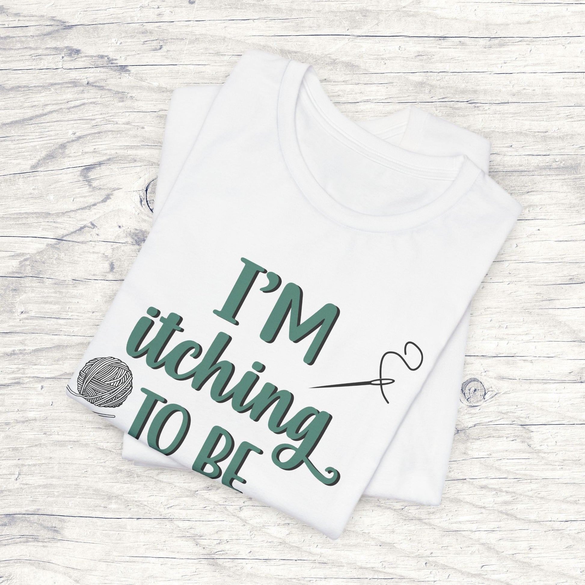 Funny Itching to be Stitching white colored folded T-Shirt with heather teal lettering, a ball of yarn, and a needle with thread