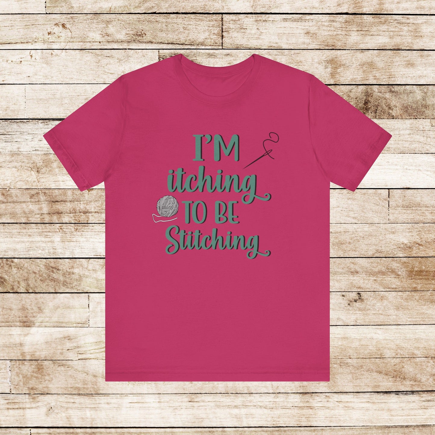 Funny Itching to be Stitching berry colored T-Shirt with heather teal lettering, a ball of yarn, and a needle with thread