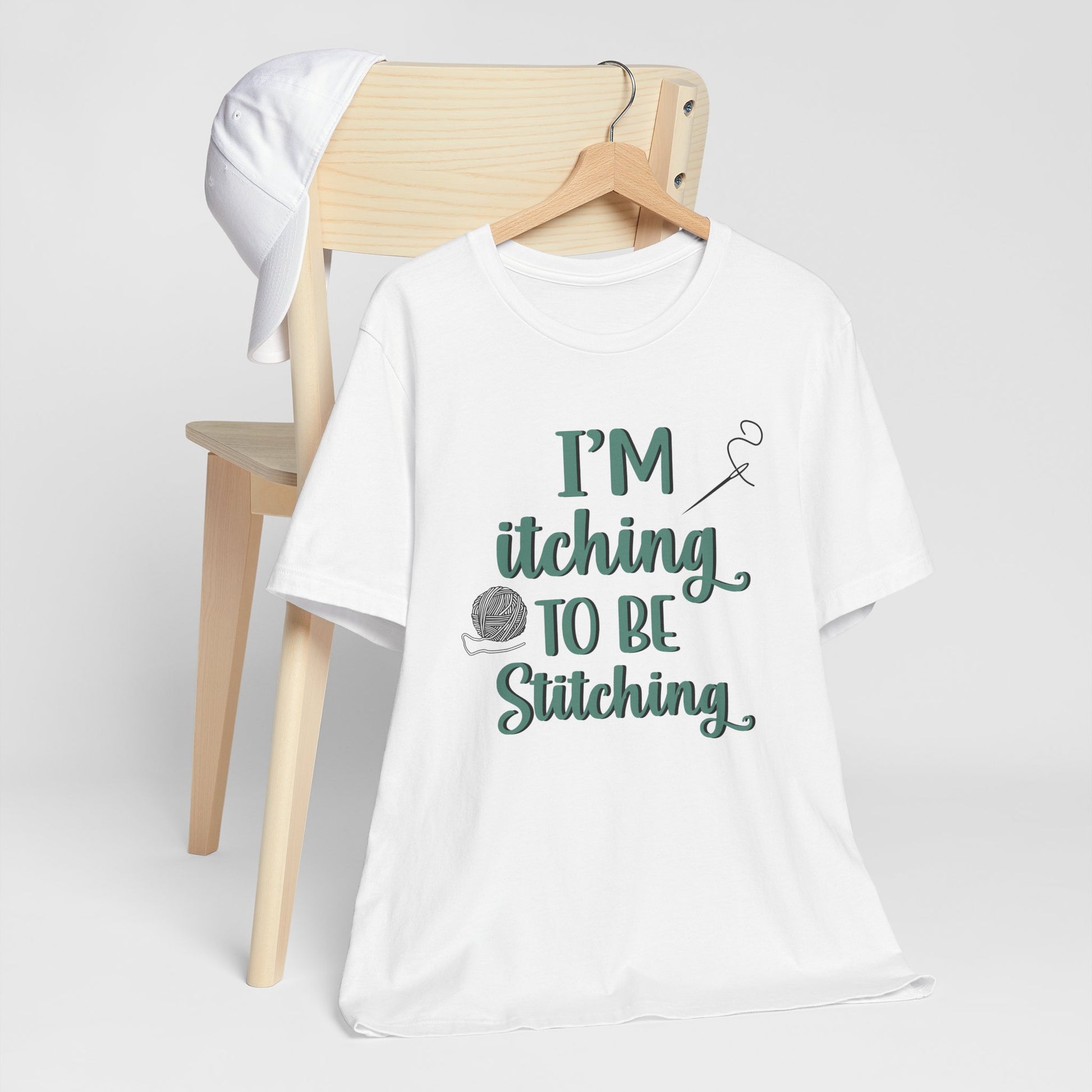 Funny Itching to be Stitching white colored T-Shirt with heather teal lettering, a ball of yarn, and a needle with thread