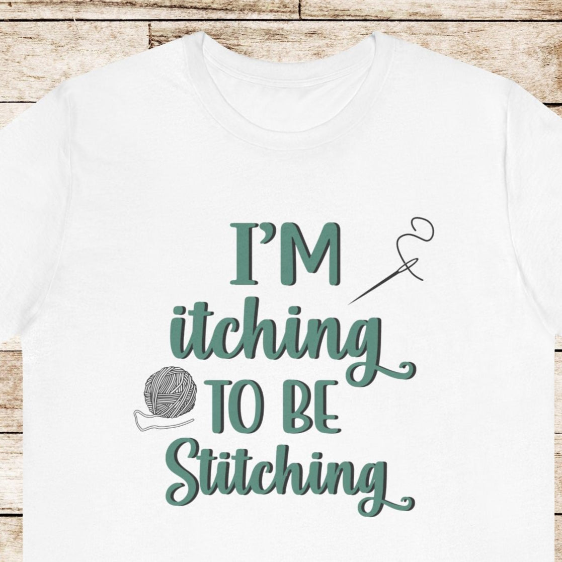 Funny Itching to be Stitching white colored T-Shirt with heather teal lettering, a ball of yarn, and a needle with thread