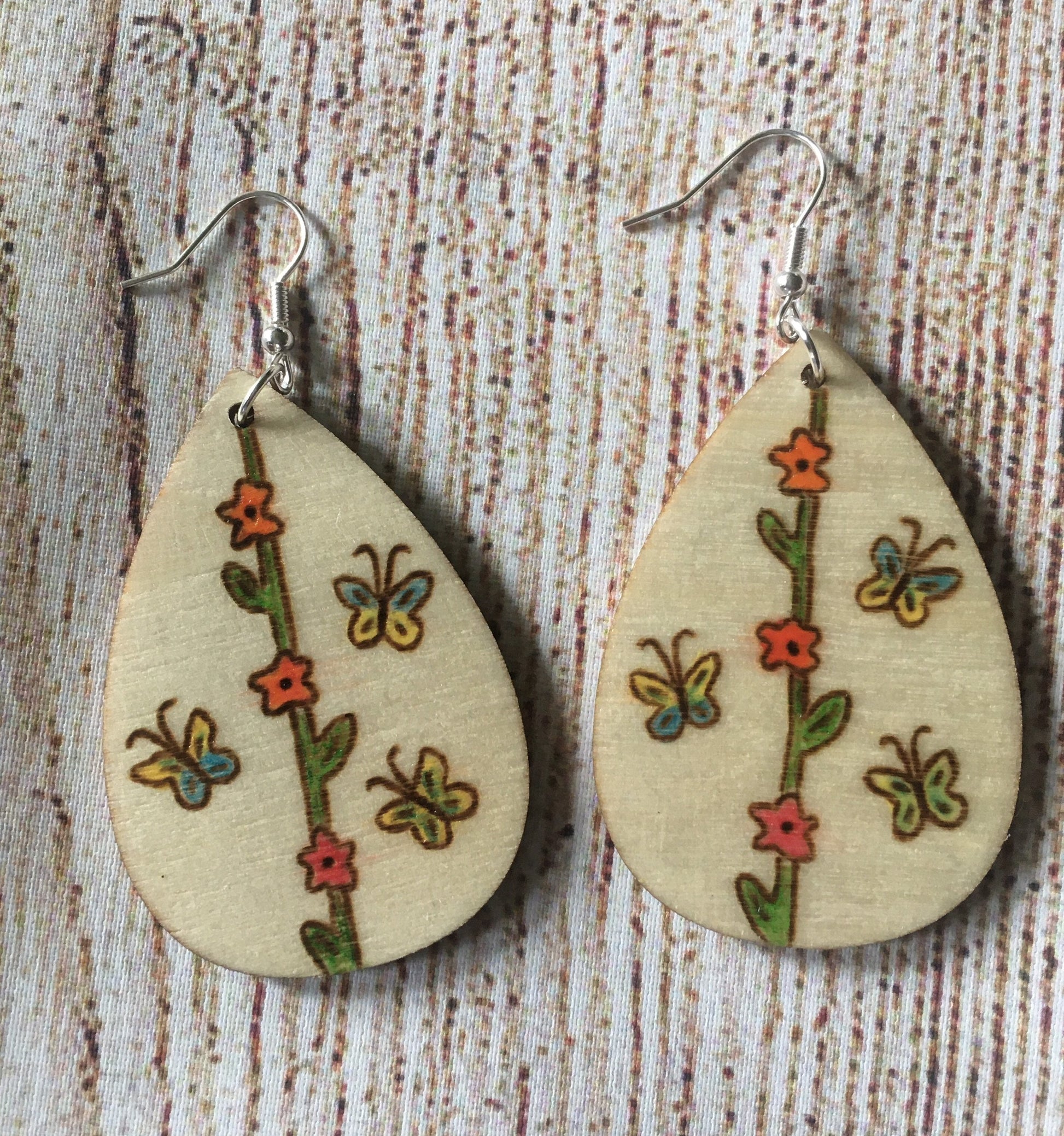 Wood burned earrings with flower vine in the middle from the top to the bottom of the earrings.  red flowers are on the vine with yellow and blue butterflies on either side. Earrings are tear drop shaped with fishhooks.