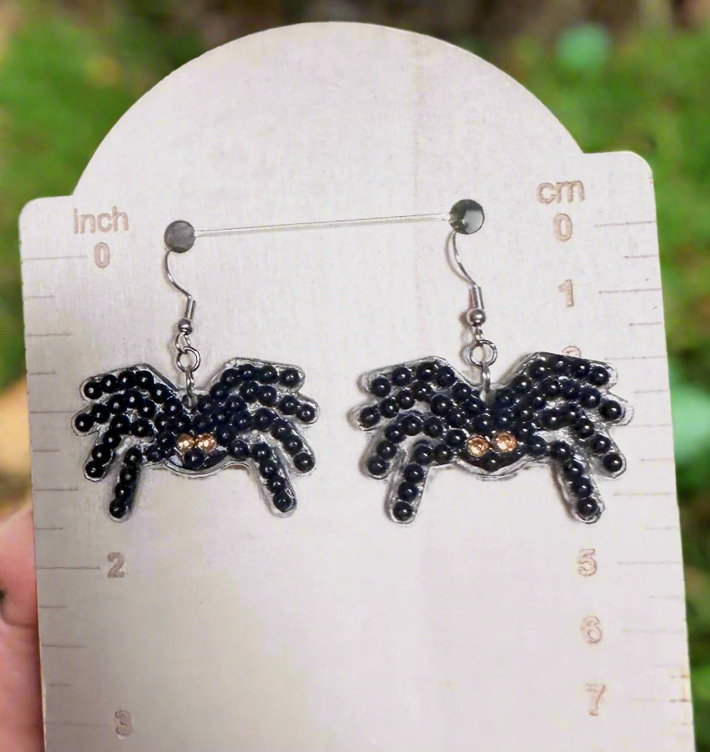 Spider-shaped earrings on a white card with measurements, against a blurred green background