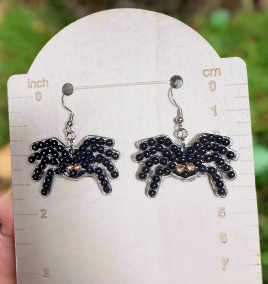 Spider-shaped earrings on a white card with measurements, against a blurred green background