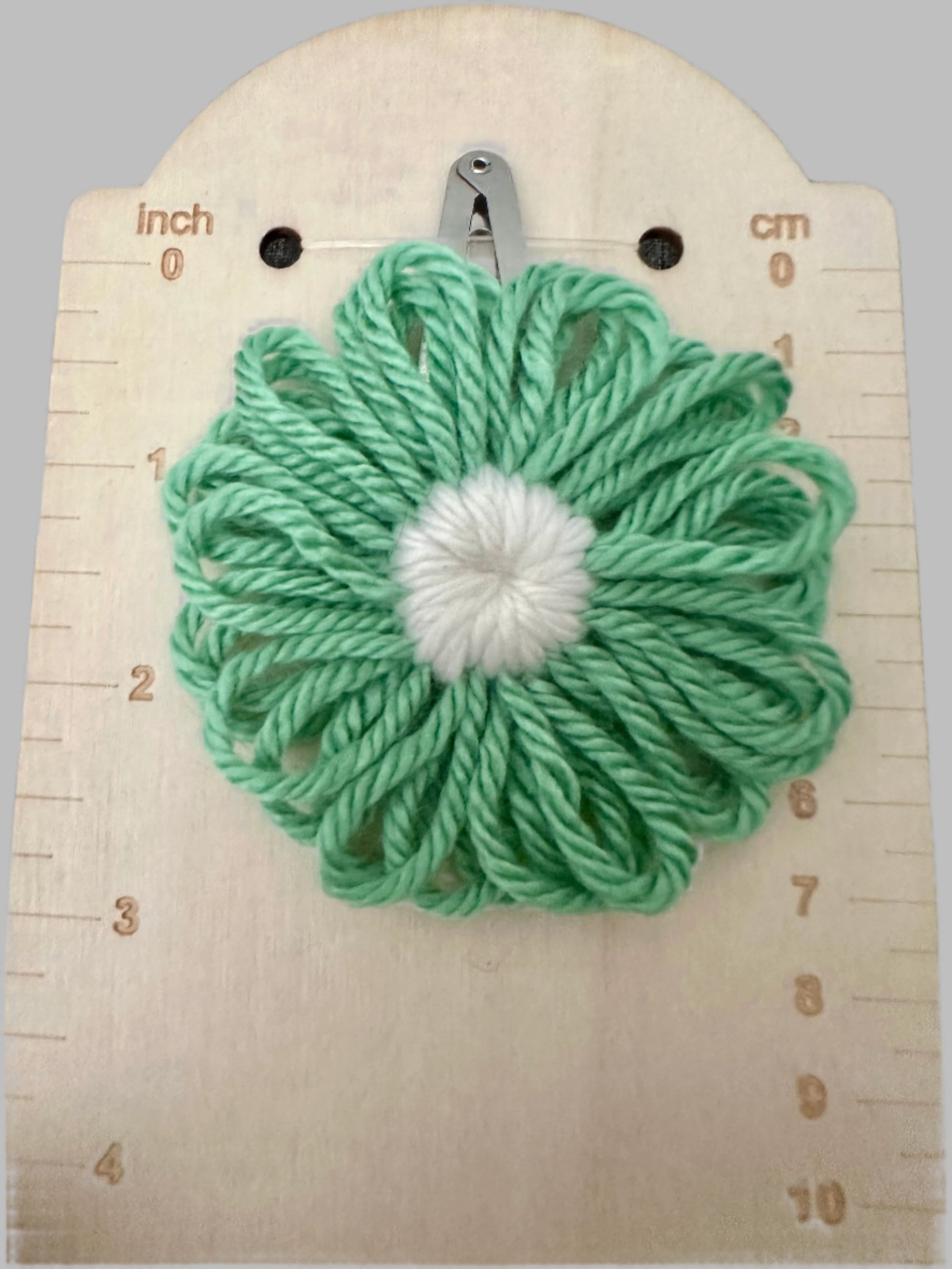 Green yarn flower with a white center on a wooden surface with measurement markings.