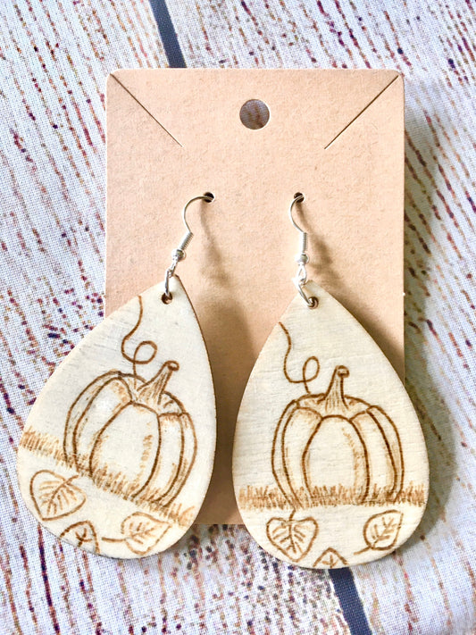 Wood burned earrings with wood burned pumpkin sitting in the grass with leaves on the ground. Earrings are tear drop shaped with fishhooks.