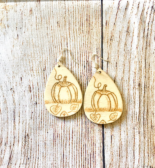 Wood burned earrings with wood burned pumpkin sitting in the grass with leaves on the ground. Earrings are tear drop shaped with fishhooks.