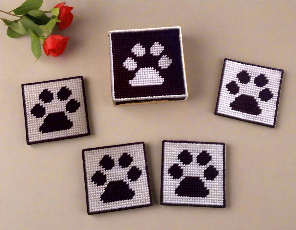 Set of four coaster with a box to hold them. Coasters have a dark brown paw print stitched with yarn on a white stitched background. The box has a white stitched pawprint on and dark brown background. All stitched with yarn on plastic canvas. 